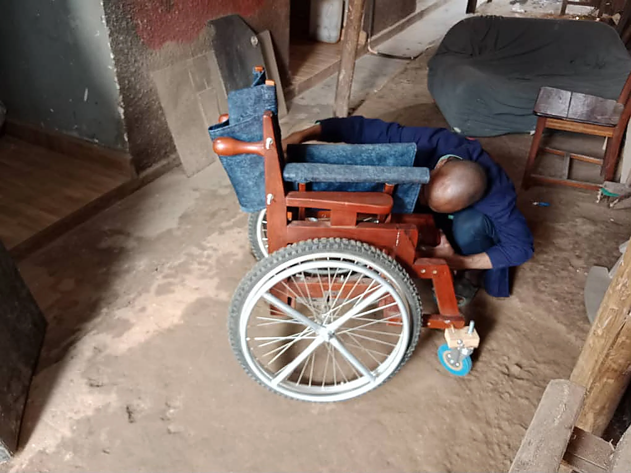 A carpenter kneeling to fit a wheel on a wooden wheelchair inside the workshop.