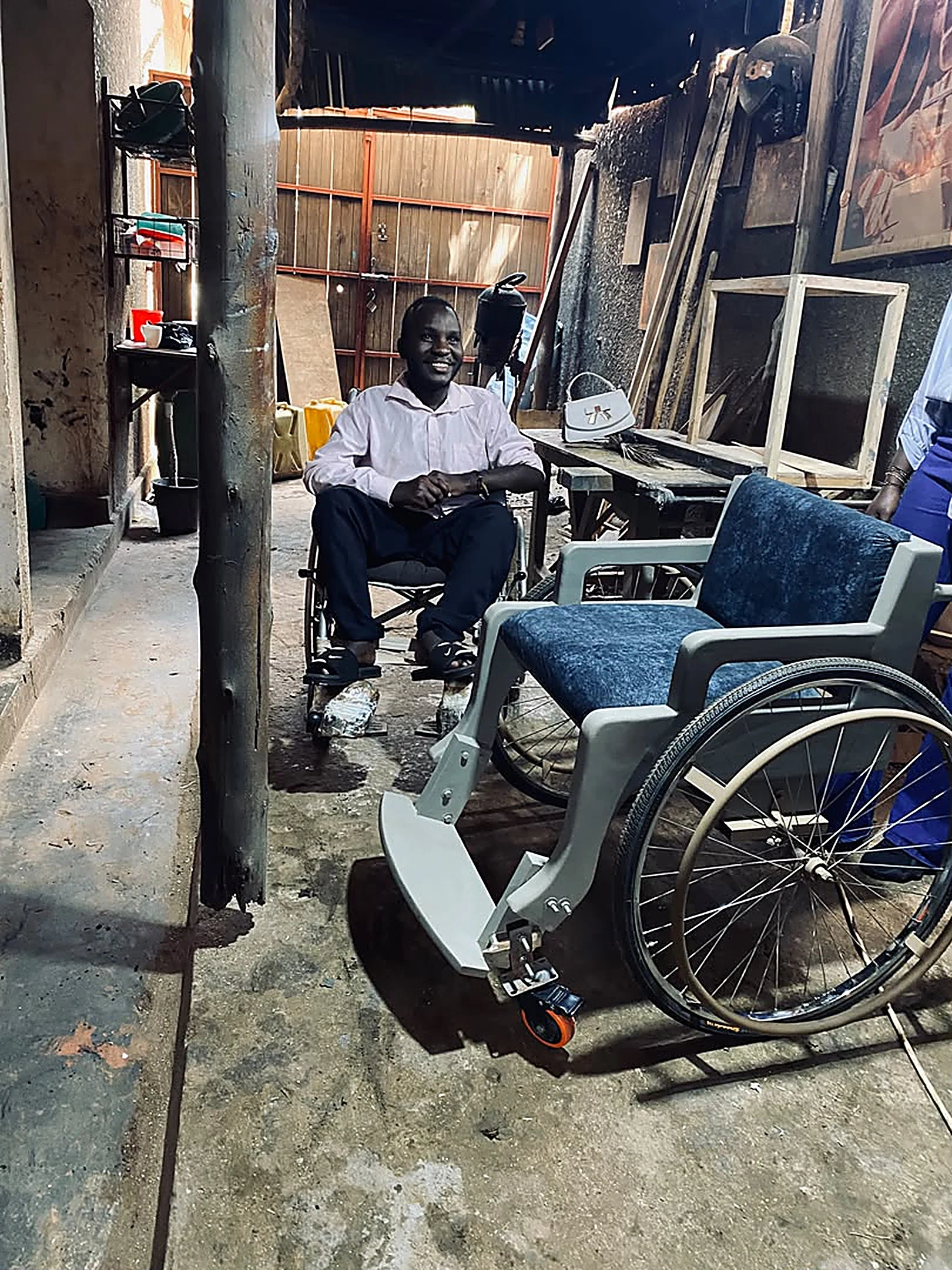 Rashid smiling in a wheelchair next to a finished terrain wheelchair in the workshop.